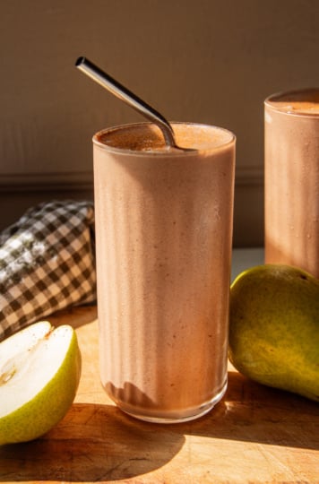A head-on shot shows a chocolate pear smoothie on a wooden board in direct sun light. A stainless straw sticks out of the smoothie and whole pears are seen nearby.