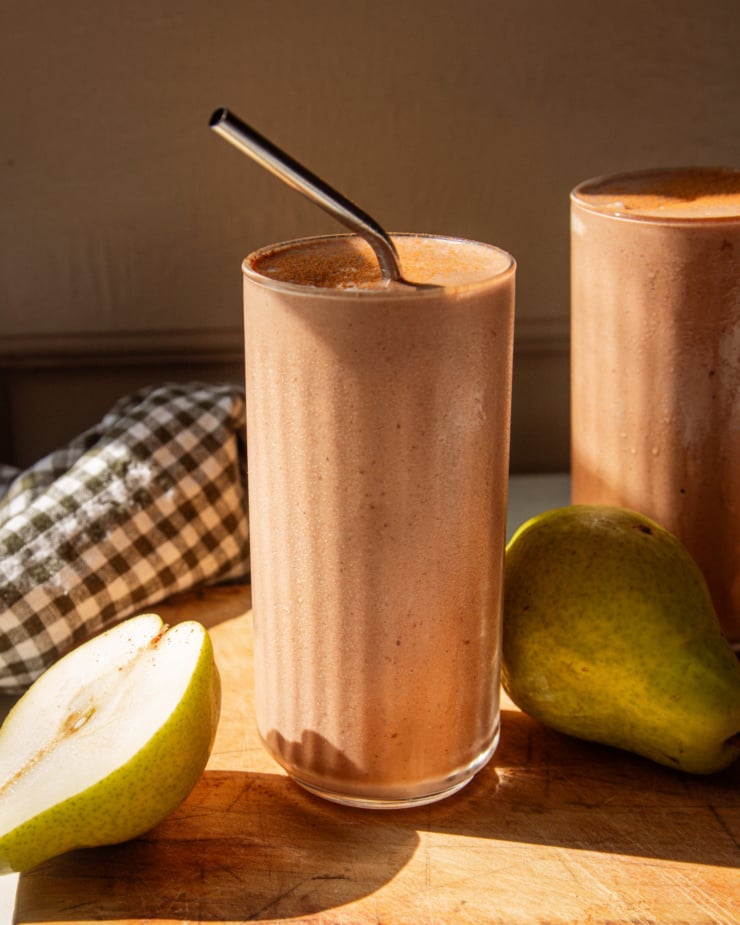 A head-on shot shows a chocolate pear smoothie on a wooden board in direct sun light. A stainless straw sticks out of the smoothie and whole pears are seen nearby.