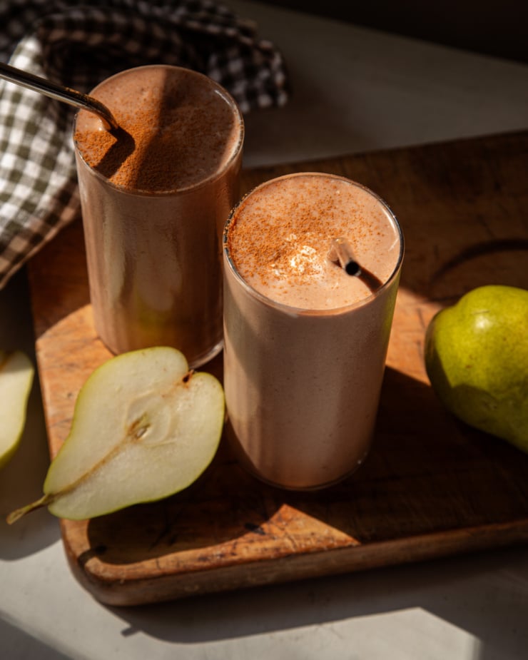 The overhead lens shows in bright sunlight, showing two chocolate pear smoothies on the wooden board. Their cinnamon sprinkled over the top, and the stainless steel straw sticks out of the glasses.