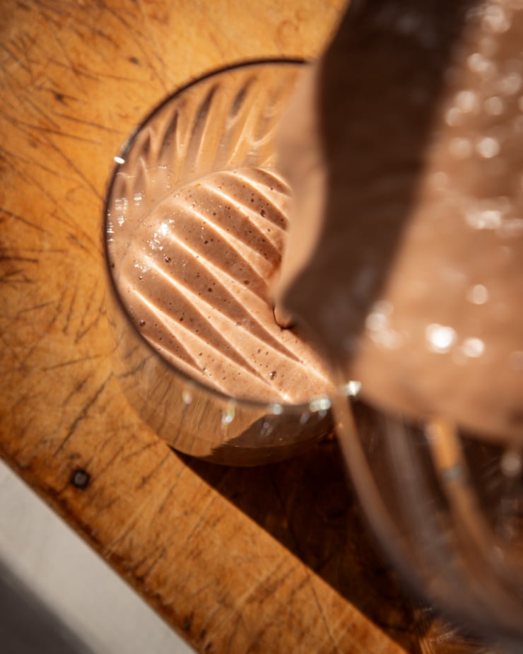 Overhead lens shows the chocolate smoothie pouring into a grooved glass. This photo was taken in direct sunlight, with the grooved texture of the glass showing stripes on the top of the smoothie surface.