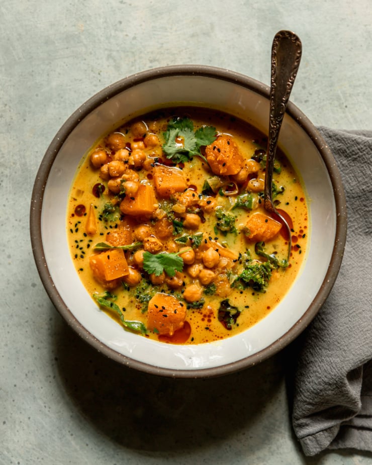 An overhead shot shows a bowl of butternut squash stew with coconut milk, kale and chickpeas. The broth is creamy and golden with bits of chopped cilantro garnishing the top. A spoon is sticking out of the bowl.