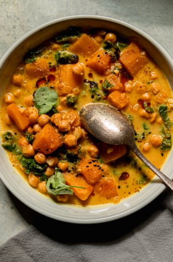 An overhead shot shows a bowl of butternut squash stew with coconut milk, kale and chickpeas. The broth is creamy and golden with bits of chopped cilantro garnishing the top. A spoon is sticking out of the bowl. This portion is also garnished with some chili crisp.