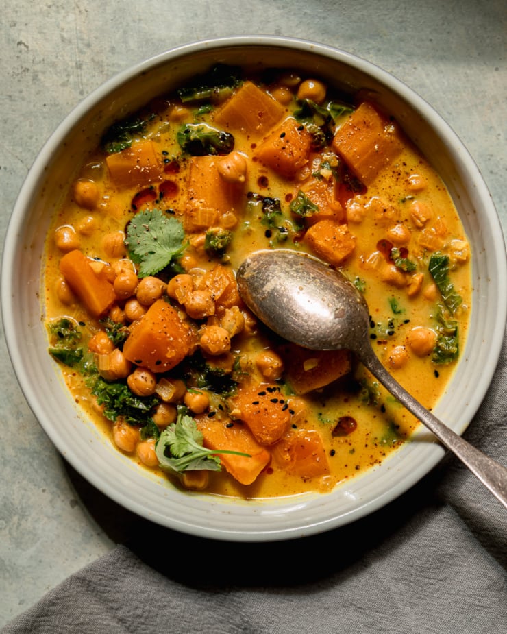 Overhead shot shows a bowl of butternut squash stewed coconut milk, kale and chickpeas. The broth is creamy golden with chopped cilantro topped with cilantro garnished. The spoon sticks out of the bowl. This part is also decorated with some chili crispy.