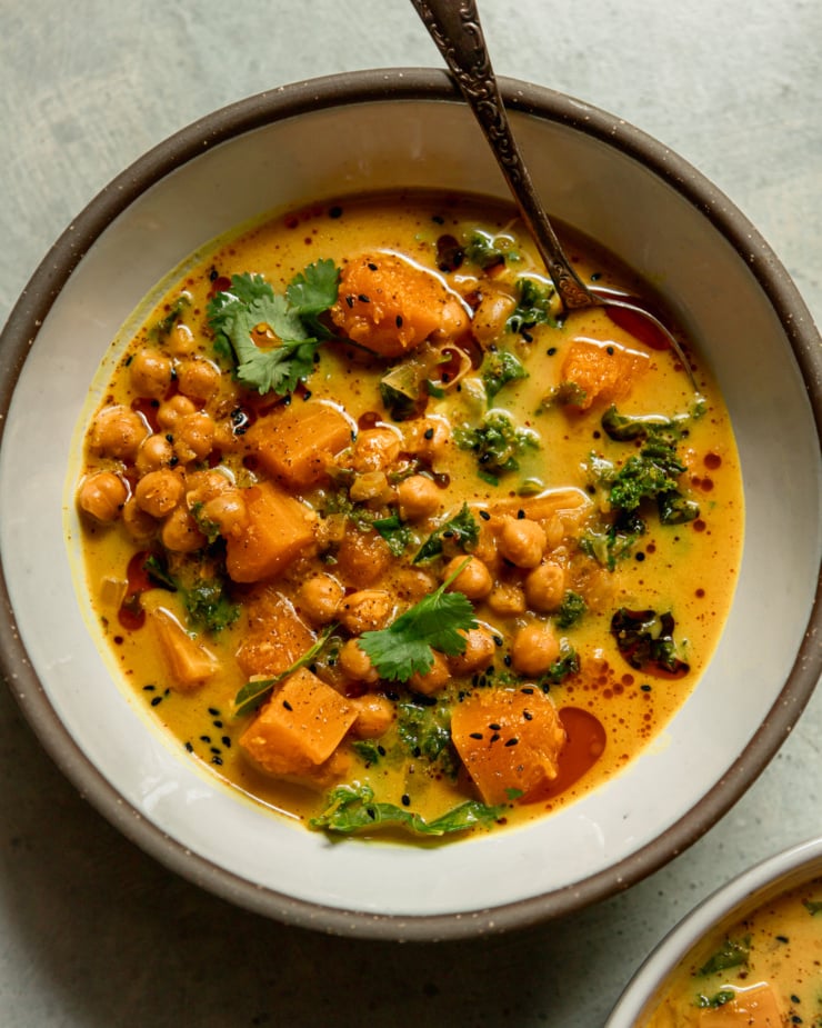 A close-up overhead shot shows a bowl of butternut squash stewed coconut milk, kale and chickpeas. The broth is creamy golden with chopped cilantro topped with cilantro garnished. The spoon sticks out of the bowl.