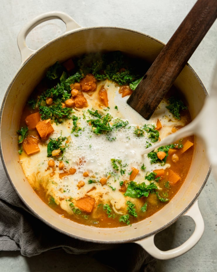 Overhead shot shows coconut milk being poured into a wooden kale, sliced, chickpeas and broth. Wooden stirring utensils stick out of the pot.