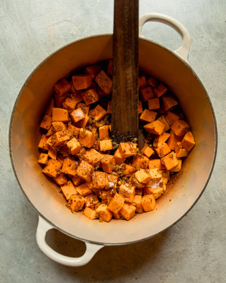Overhead shot shows the pumpkin cubes sautéed in a large pot with spices. Wooden stirring utensils stick out of the pot.