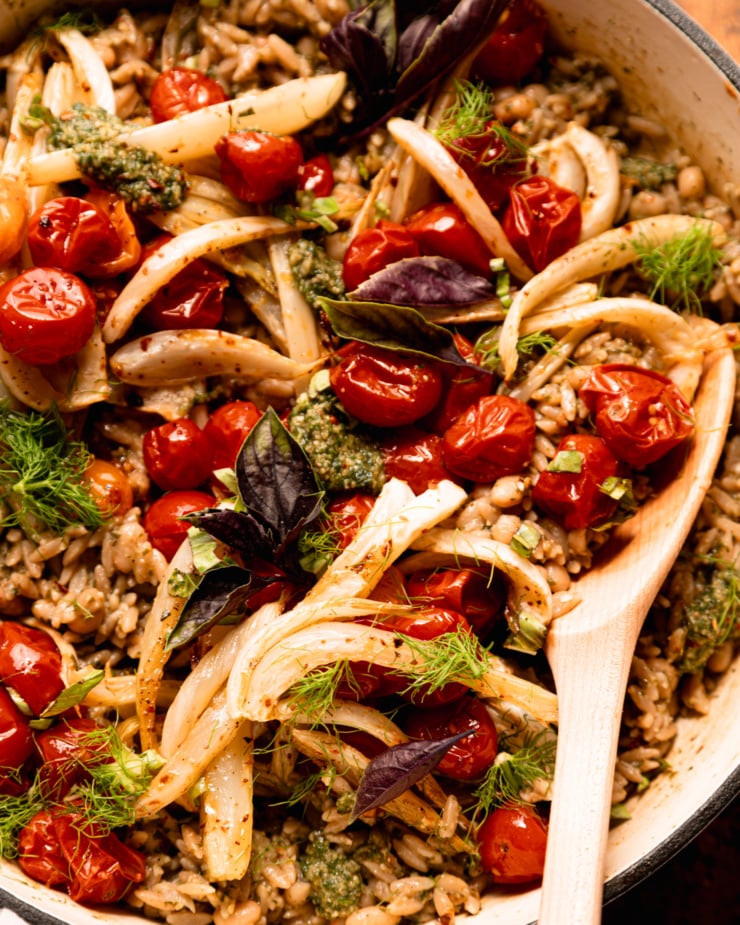 A close-up overhead shot shows the Orzo dish of pesto, topped with roasted cherry tomatoes, fennel, fresh basil and fennel leaves. The wooden spoon is sticking out everything contained on the siding.