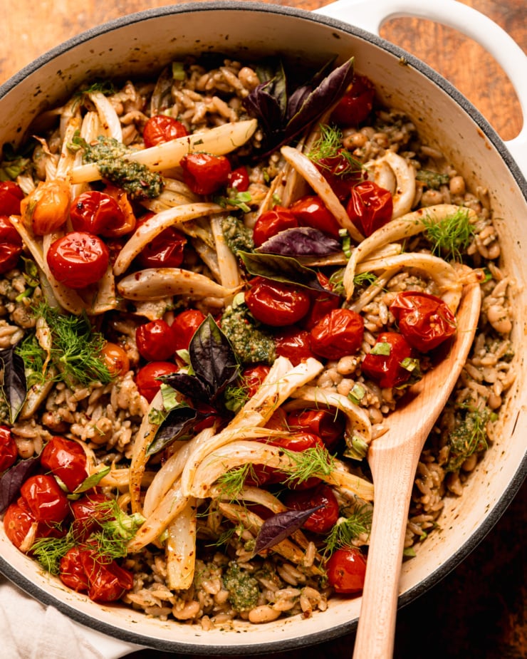 A close-up overhead shot shows the Orzo dish of pesto, topped with roasted cherry tomatoes, fennel, fresh basil and fennel leaves. The wooden spoon is sticking out everything contained on the siding.