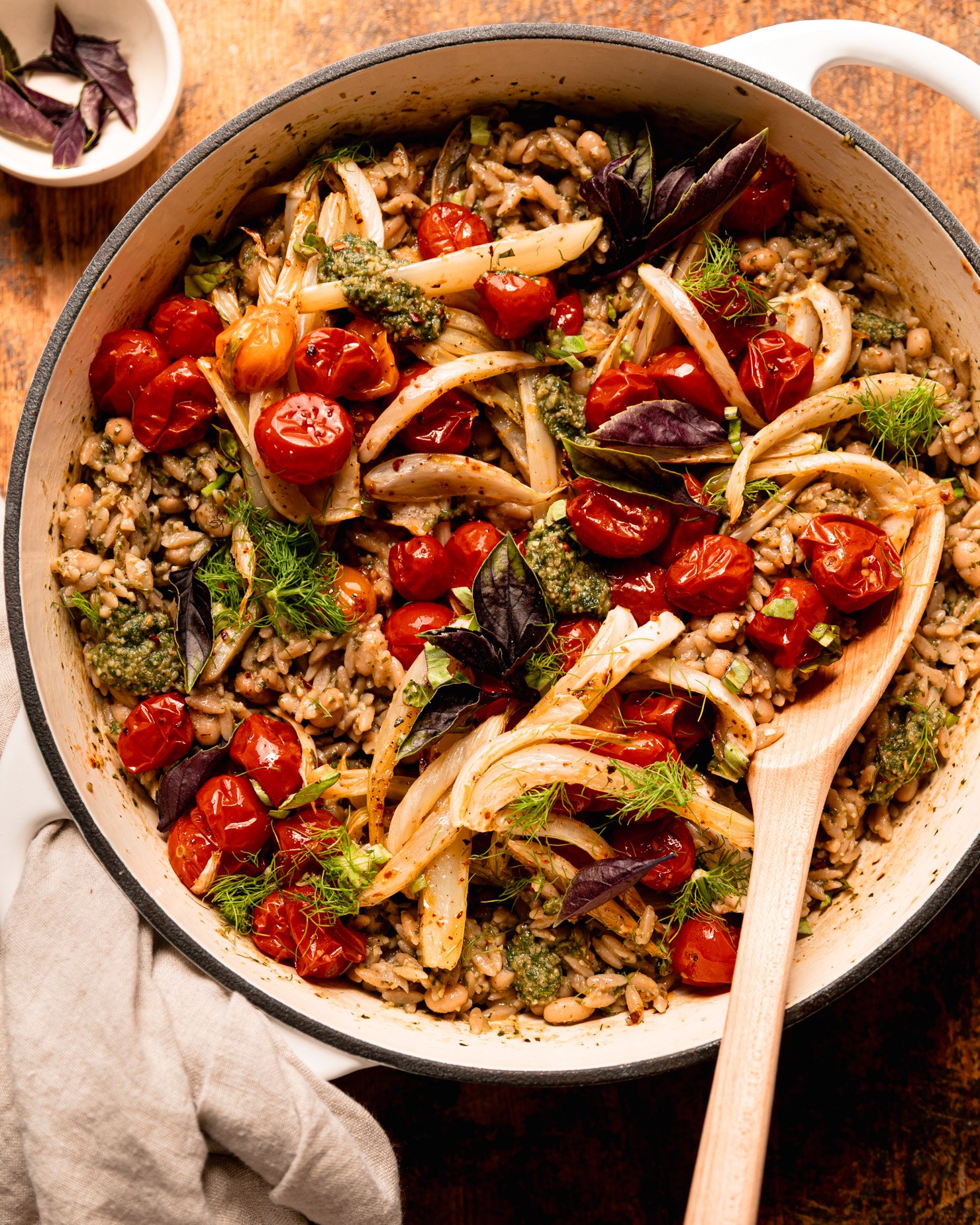 The overhead shot shows the Orzo dish of pesto, topped with roasted cherry tomatoes, fennel, fresh basil and fennel leaves. The wooden spoon is sticking out everything contained on the siding.