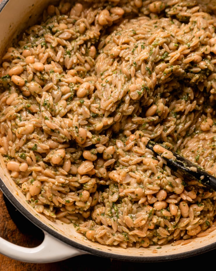 Overhead shot shows a pot filled with cooked Orzo pasta, white beans and pesto sauce. The texture is creamy.