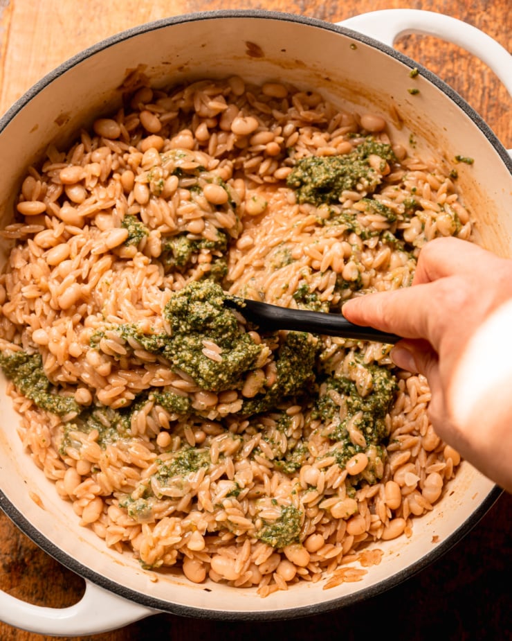 Overhead shot shows a hand stirring the garlic sauce with a spatula into cooked orzo pasta and white beans in a pot.