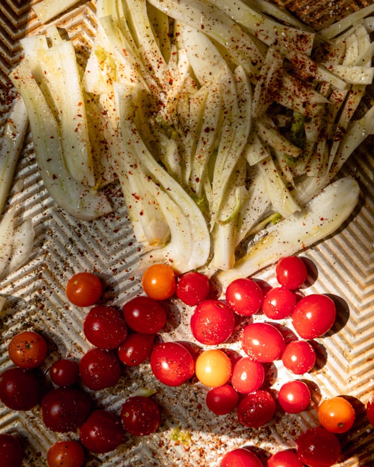 Overhead shot shows fresh fennel and cherry tomatoes thinly sliced ​​on a baking tray. They sprinkle with olive oil and season with chili flakes, salt and pepper.