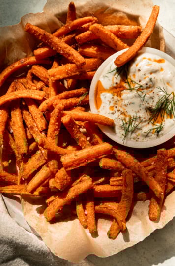 An overhead shot shows a paper-lined dish filled with roasted carrot fries. A small bowl of garlicky dill yogurt dip is seen to the side. The dip is garnished with chili oil and fresh dill. The photo is taken in direct sunlight.