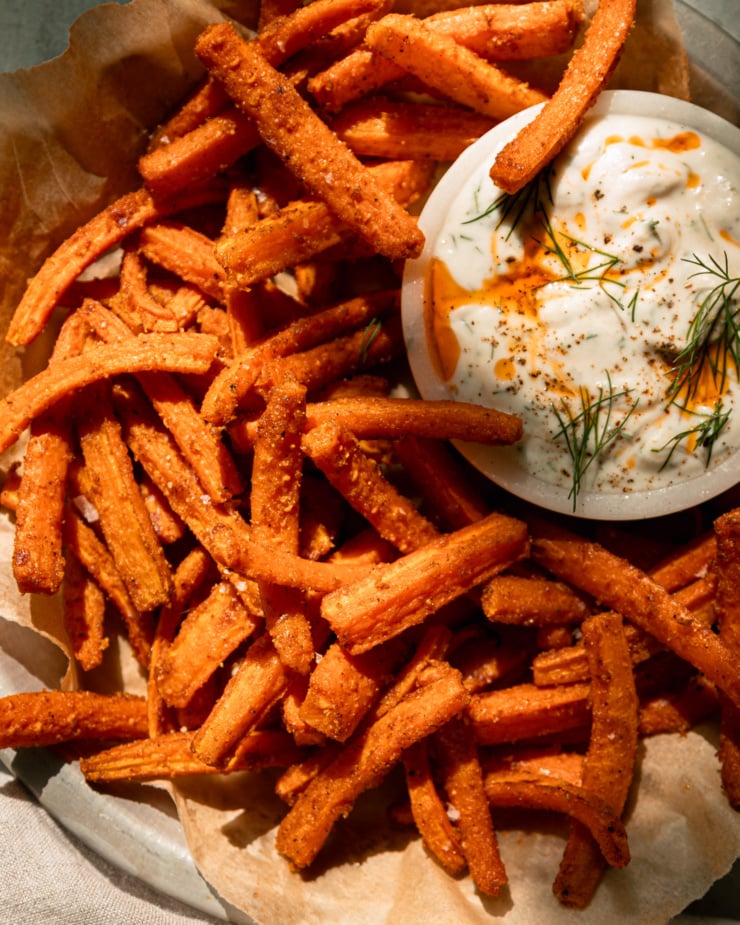 A close-up overhead shot shows a paper-lined plate filled with roasted carrot fries. A small bowl of gilded dill yogurt dipped aside. Dip in chili oil and fresh dill. This photo is in direct sunlight.