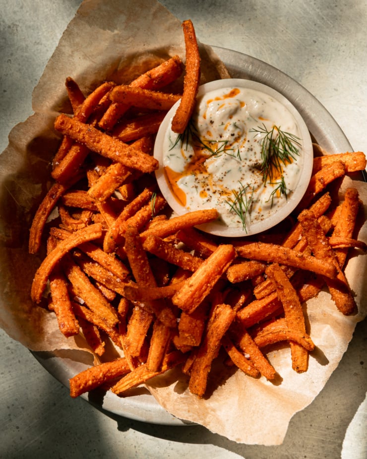 The overhead shot shows a paper-lined plate filled with roasted carrot fries. A small bowl of gilded dill yogurt dipped aside. Dip in chili oil and fresh dill. This photo is in direct sunlight.