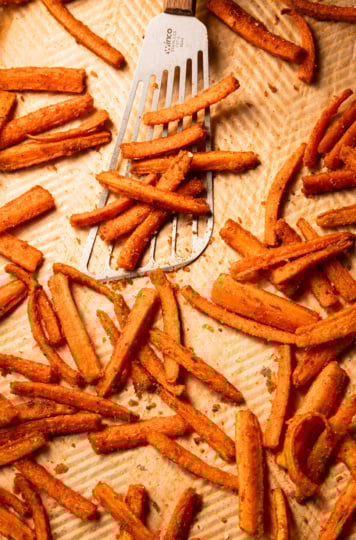 An overhead shot shows roasted carrot fries on a parchment-lined baking sheet. A fish spatula is lifting some off the baking sheet.
