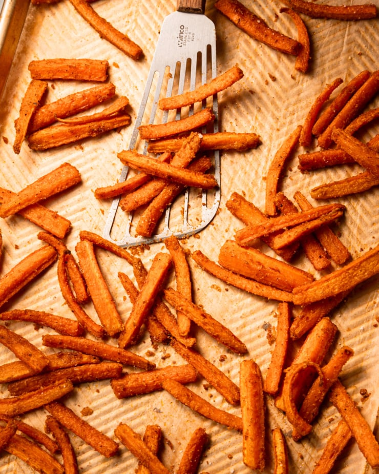 Overhead shot shows grilled carrot fries on a parchment-lined baking tray. A fish spatula will put some on the baking sheet.