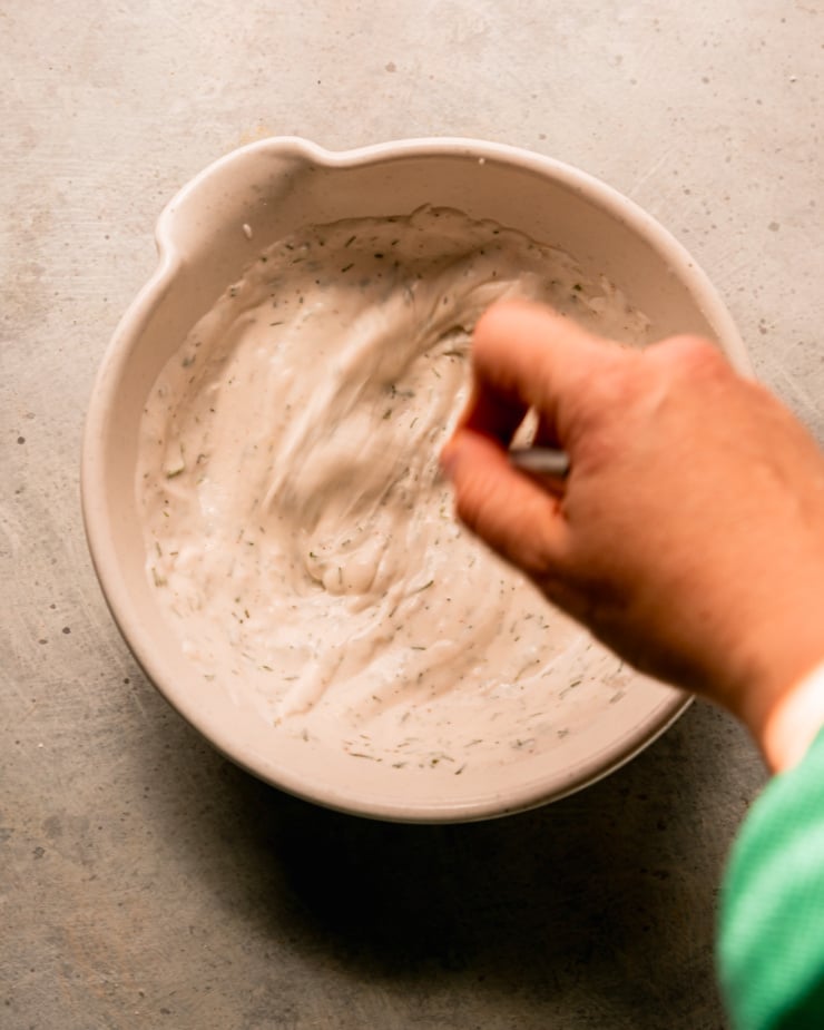 Overhead shot shows a hand stirring the sauce of yogurt together in a bowl. The stirring action is blurry.