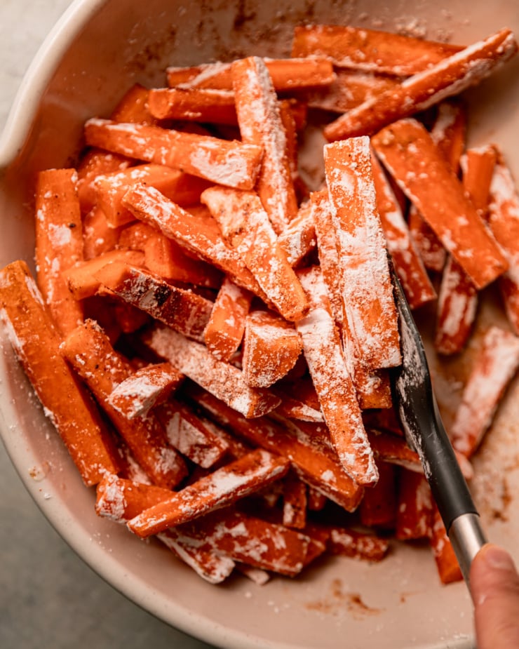 Close up overhead shot showing a hand stirring carrots in a bowl with a spatula. Carrots are coated with oil, corn starch and spices.