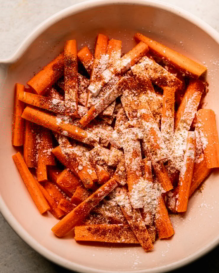 Overhead shot shows chopped carrot sticks sprinkled with cornstarch, spices, salt and pepper. All in a large ceramic bowl.