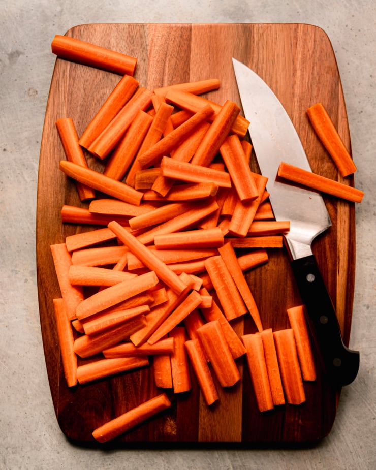 Overhead shot showing a pile of carrots cut into sticks on wooden cutting board. I saw a chef's knife.