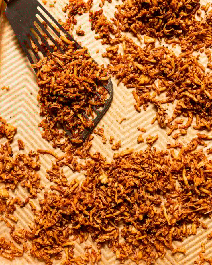 A close-up overhead shot shows a baking sheet filled with crispy and chewy shredded grilled tofu. A metal spatula can be seen nearby.