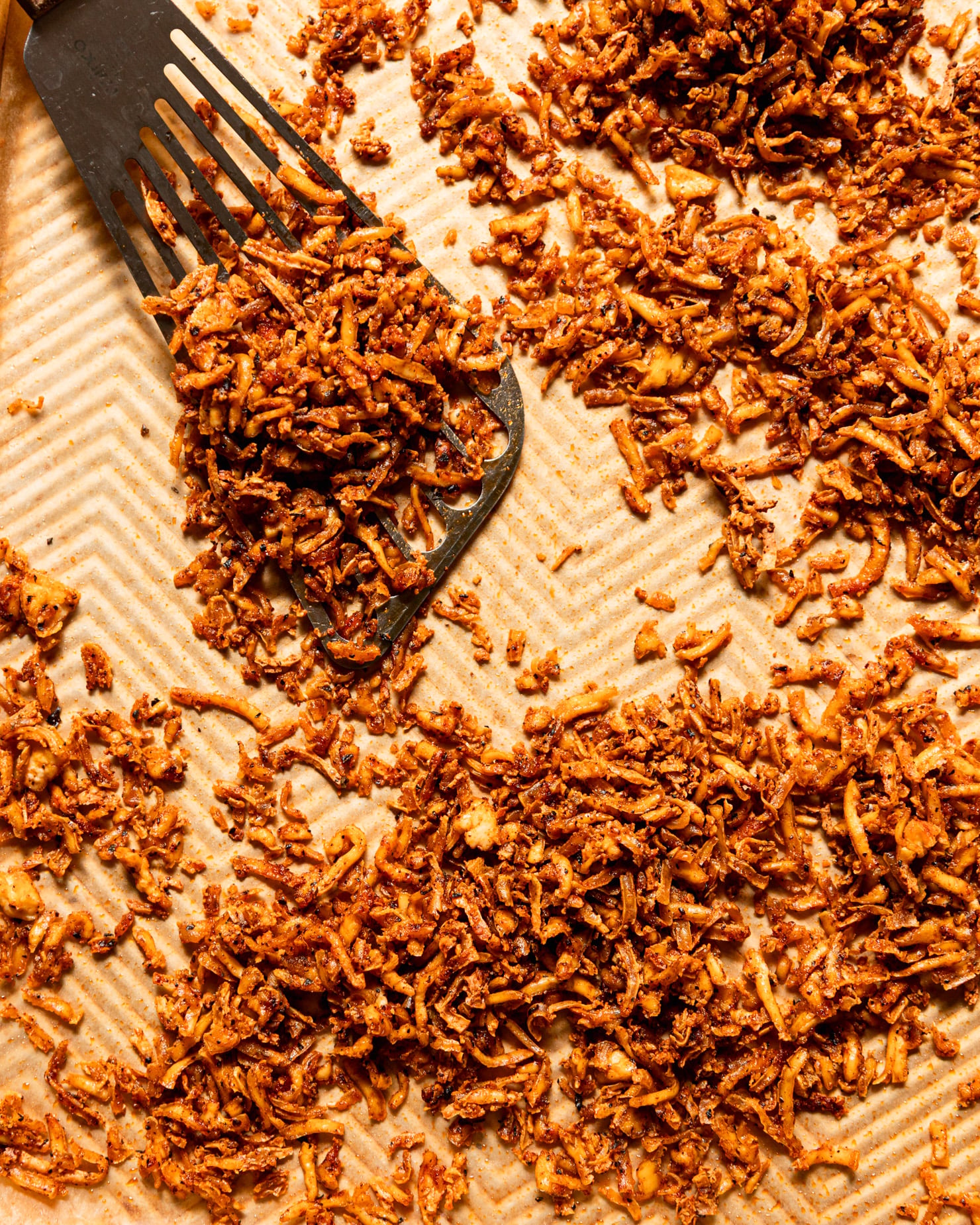 An overhead, up close shot shows a baking sheet filled with crispy and chewy baked shredded tofu. A metal spatula is seen to the side.