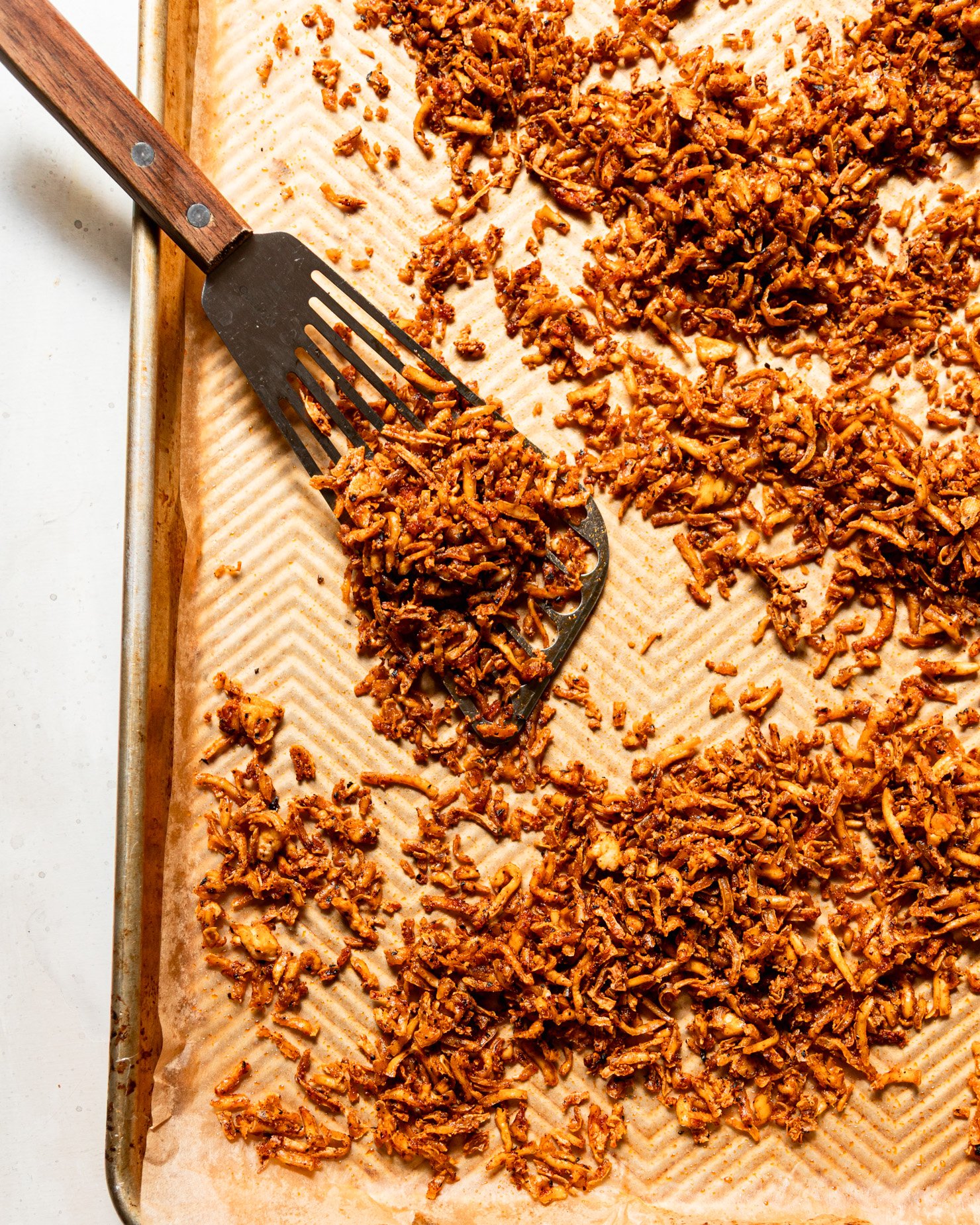 The top-down photo shows a baking sheet filled with crispy and chewy shredded grilled tofu. A metal spatula can be seen nearby.