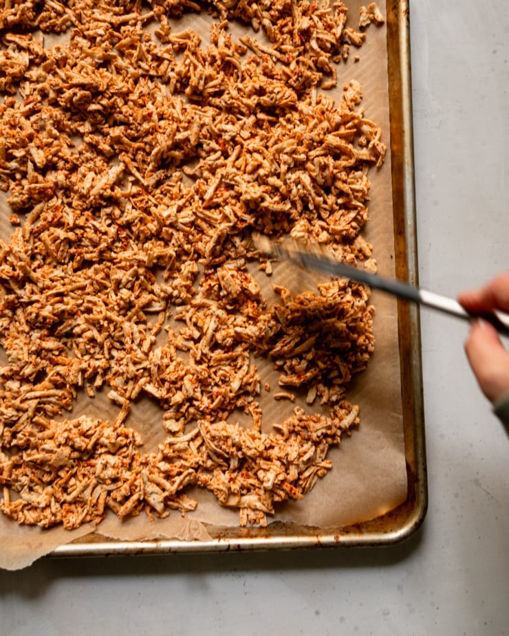 The top-down photo shows a hand using a spatula to spread chopped spiced tofu onto a parchment-lined baking sheet. There is motion blur around the spatula.