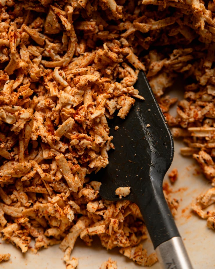 A close-up overhead shot shows shredded tofu tossed with oil, spices and cornstarch. A spatula can also be seen.