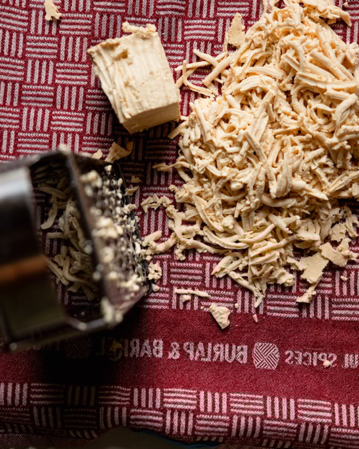 Top view shows a box grater, a block of tofu, and some shredded tofu; all placed on a clean kitchen towel.