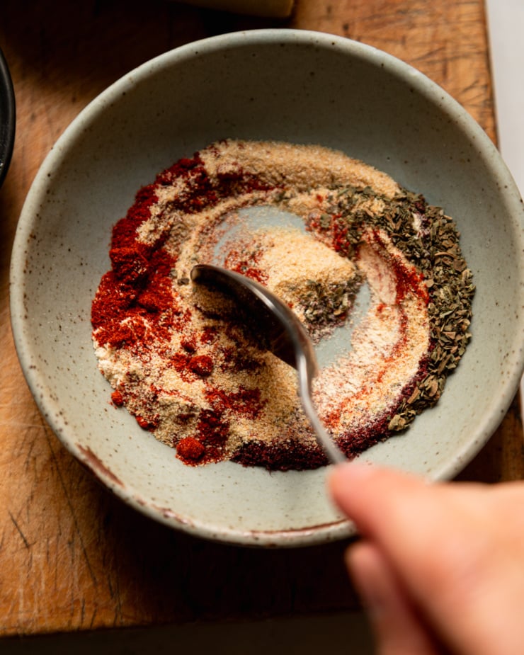 Top view photo shows a hand stirring some spices in a bowl with a spoon.