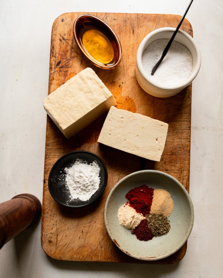An overhead shot shows a cutting board with a few items on it: a bowl of oil, a bowl of salt, a bowl of spice mix, a bowl of cornstarch, and a block of tofu.