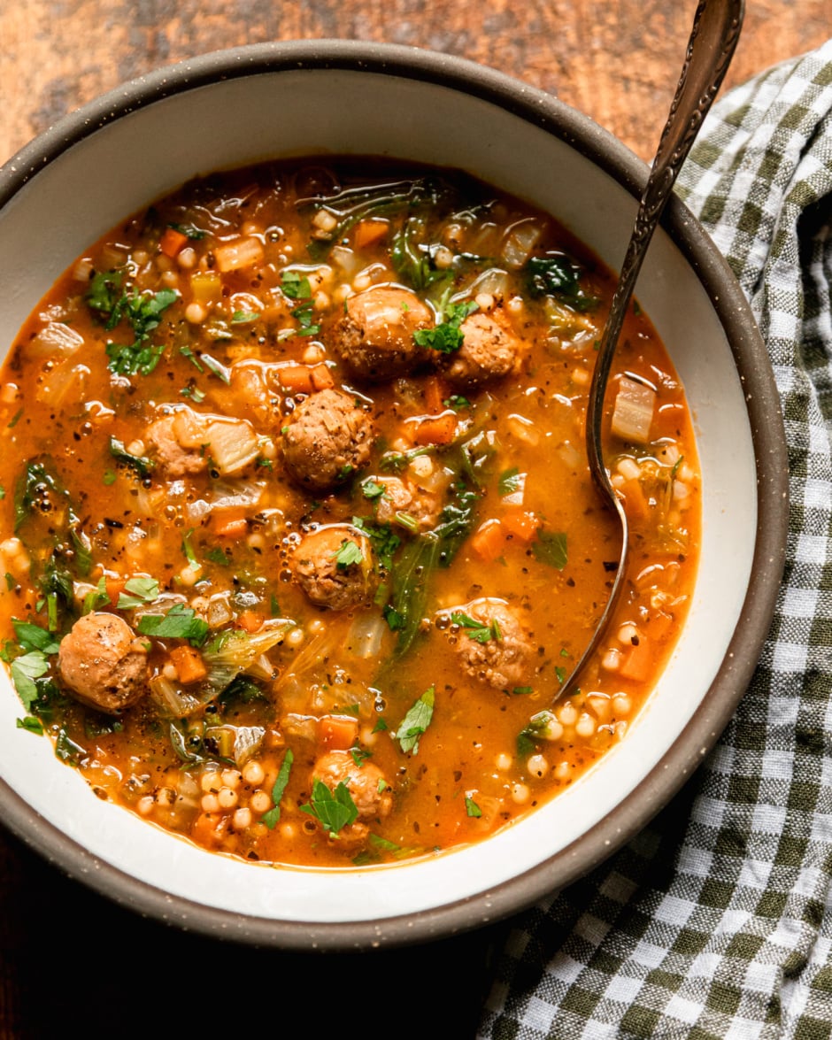 An overhead shot shows an individual bowl of vegan Italian wedding soup with little vegan sausage meatballs, tiny pasta pearls, wilted chopped escarole, and sautéed veggies. The top of the soup is garnished with chopped parsley. A spoon sticks out of the bowl and a checked napkin is nearby.