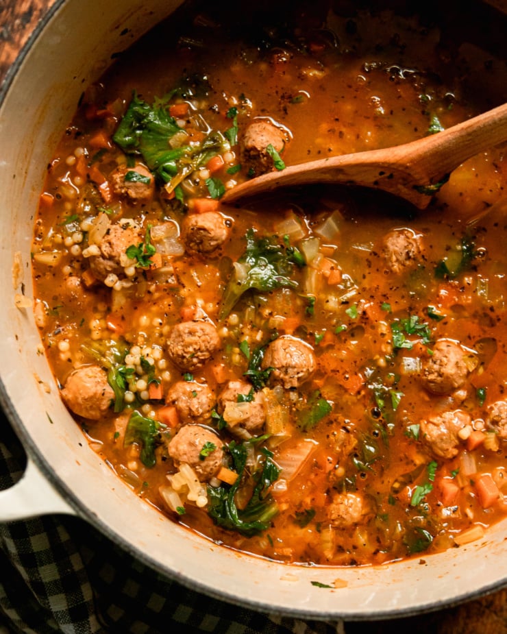 An overhead shot shows a pot of vegetarian Italian wedding soup. You'll see wilted scraps, tiny pasta dishes, and vegan sausage meatballs floating above the ground. The soup is garnished with chopped parsley and a wooden spoon can be seen dipping into the soup.