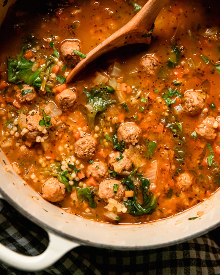 A close overhead shot shows a pot of vegetarian Italian wedding soup. You'll see wilted scraps, tiny pasta dishes, and vegan sausage meatballs floating above the ground. The soup is garnished with chopped parsley and a wooden spoon can be seen dipping into the soup.