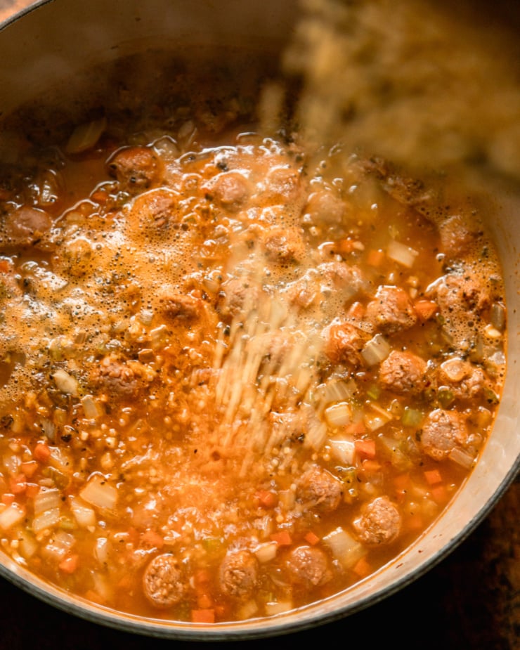 An overhead shot shows small pasta being poured into a pot of boiling soup.
