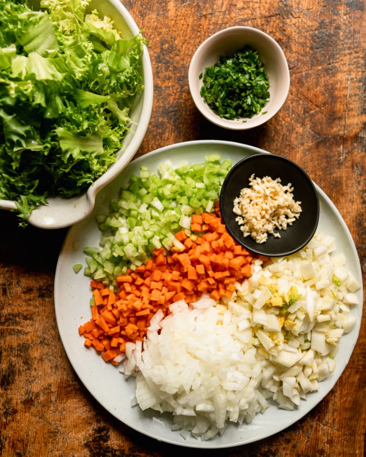 An overhead shot shows a bowl of chopped grounds, a small bowl of chopped parsley, a small bowl of chopped garlic and a dinner plate with chopped fennel, onions, carrots and celery.