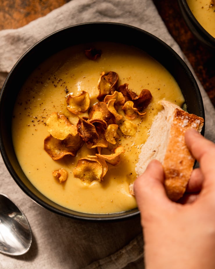 An overhead shot shows a hand dipping a crust of bread into a bowl of creamy vegan parsnip soup. The soup is garnished with parsnip chips.