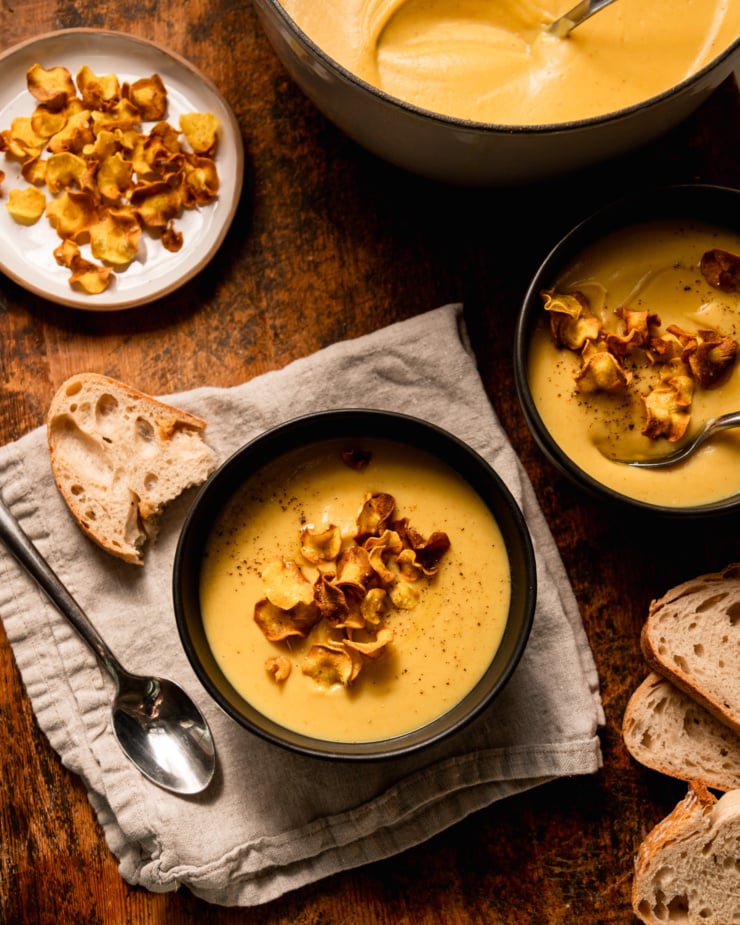 The overhead photo shows two individual bowls of parsnip soup, garnished with parsnip slices. A plate of parsnips and a pot of soup were nearby. Slices of bread can also be seen.