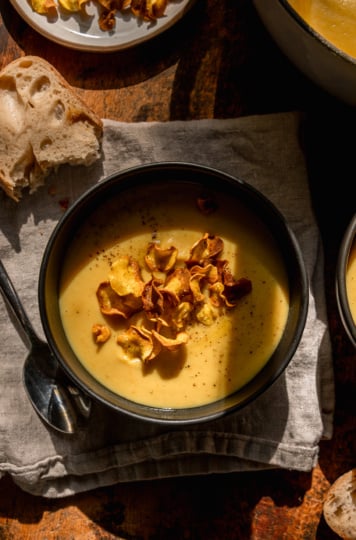 An overhead shot shows an individual serving of parsnip soup garnished with parsnip chips. Bread is nearby and the photo is taken in direct sunlight.