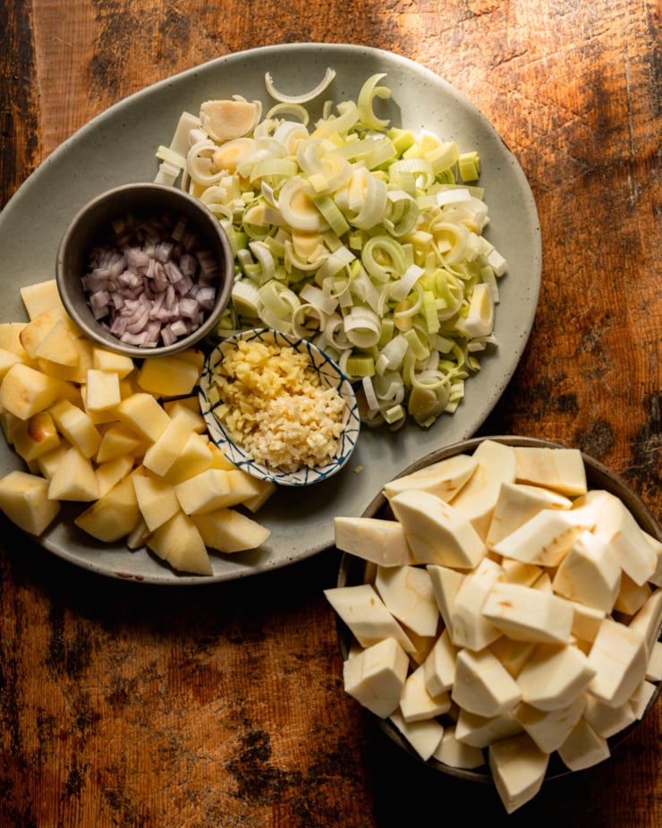 Top view photo showing chopped leeks, chopped shallots, chopped apple, chopped ginger, chopped ginger and chopped parsnips; all on their own plate/bowl as part of soup preparation.