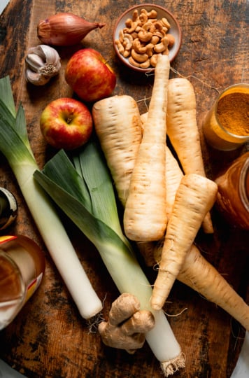 An overhead shot shows ingredients needed for a hearty vegan soup, all on a worn wood cutting board.