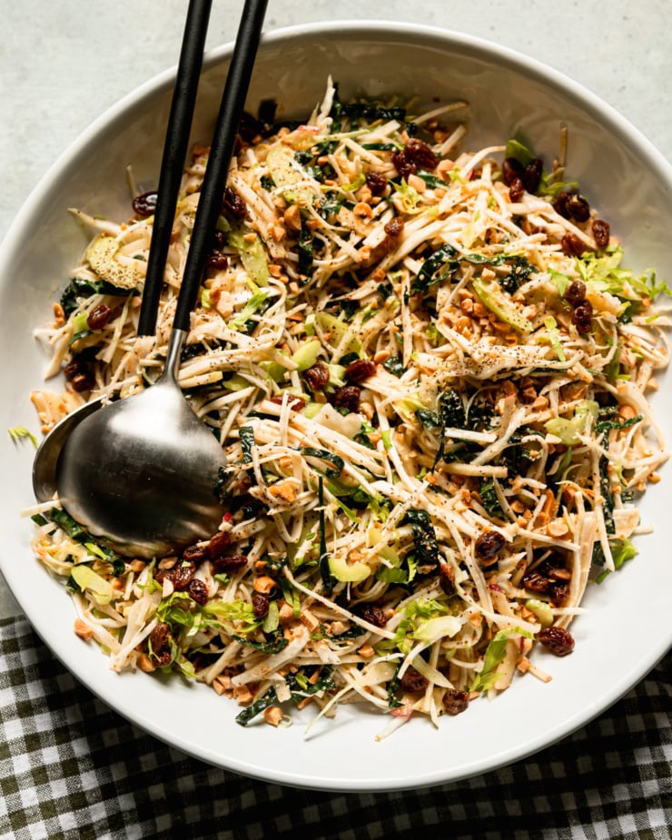 Top view shows celery root salad in a wide bowl. The salad also features celery, kale, cabbage, apples, peanuts and raisins. Shred all fruits and vegetables. There was a plaid napkin next to it and tongs inside the salad.
