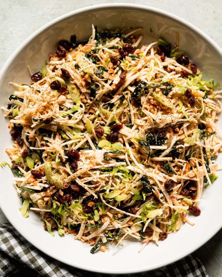 An up close, overhead shot shows a celery root salad in a wide bowl. The salad also features celery, kale, cabbage, apples, peanuts and raisins. All of the fruit and vegetables are julienned fine.