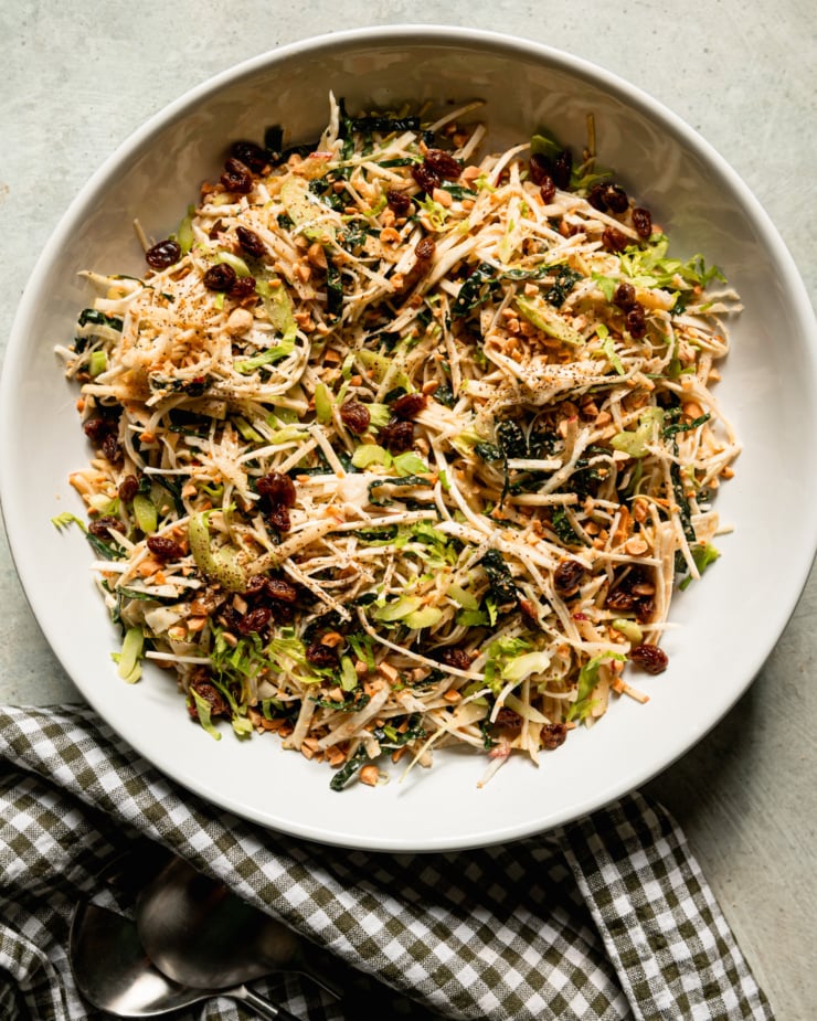 Top view shows celery root salad in a wide bowl. The salad also features celery, kale, cabbage, apples, peanuts and raisins. Shred all fruits and vegetables. There is a plaid napkin next to it.