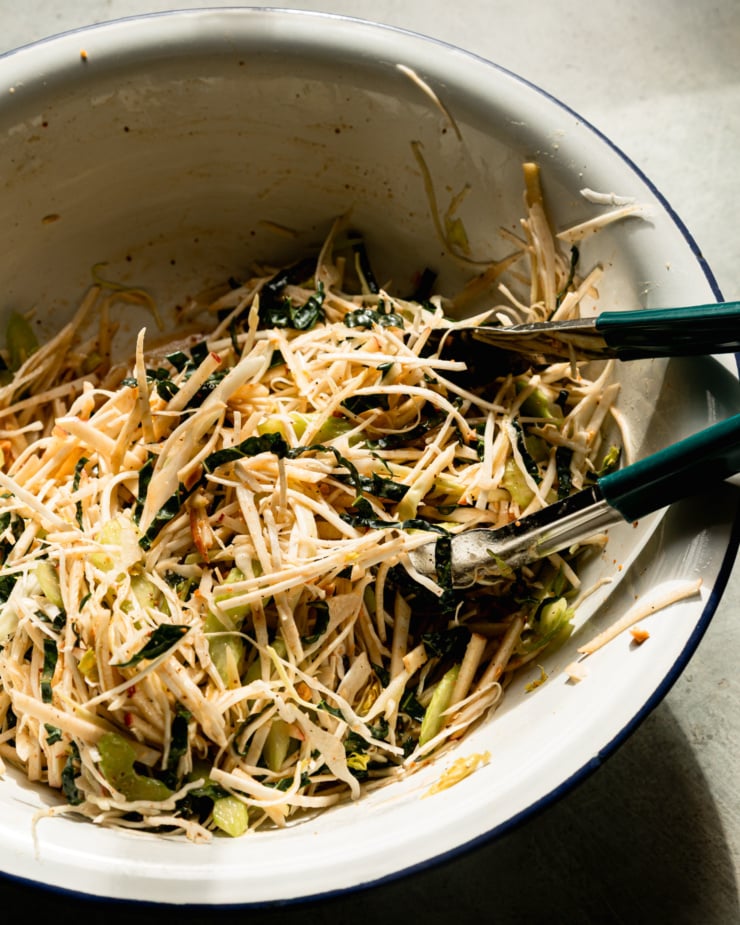 Slight 3/4 angle image shows a salad-like salad just served in an enamel bowl. The tongs hang from the side of the bowl.
