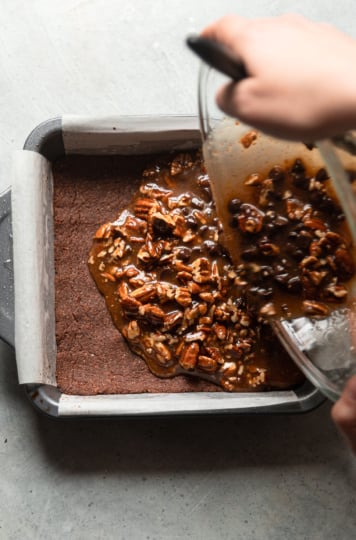 An overhead shot shows a pair of hands pouring a gooey pecan chocolate filling on top of a pan with a baked chocolate crust.
