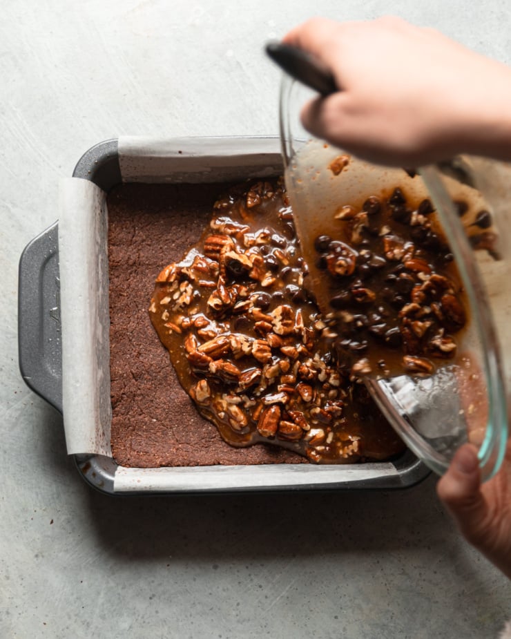 The overhead photo shows a pair of hands pouring the gooey pecan-chocolate filling onto a pan with a toasted chocolate crust.