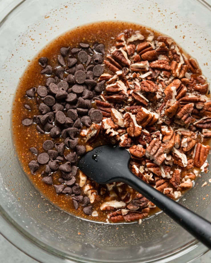 An overhead view shows a gooey pecan bar in a bowl, topped with chopped pecans and chocolate chips, about to be folded in.